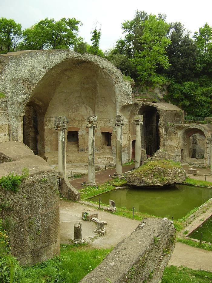 An image of stone ruins is shown. In the background a line of tall, green trees is seen on a pale blue and white sky. In front of the trees on the left, half of a domed stone structure stands – exposing the inside. Windows can be seen and four tall columns stand in front. A brick wall is shown in the left forefront corner with green branches sticking out the top. In front of the four columns a square area filled with green colored water and bordered by green moss is shown with a large rock overhanging on the top corner. Behind the water two tall rounded arches can be seen and fenced walkways behind the arches. Another area with water can be seen cut off on the right and a stone walkway is seen in the forefront.