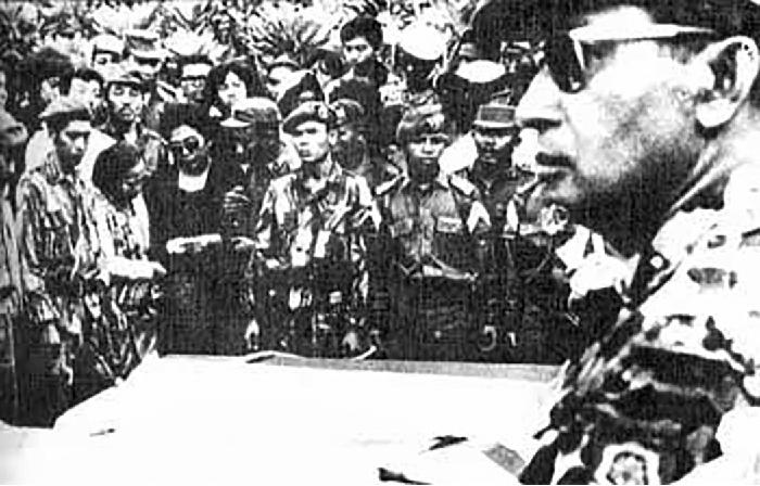 A photograph of the profile of a man in military gear is shown. He is in the front right of the picture and has a cap on, sunglasses, and an emblem on his left shoulder. There is a flat square platform in front of him and group of people in the background. In the group, a woman dressed in a black dress and sunglasses stands, surrounded by many others Some of the men are dressed in military gear with caps and other women are in black dresses.