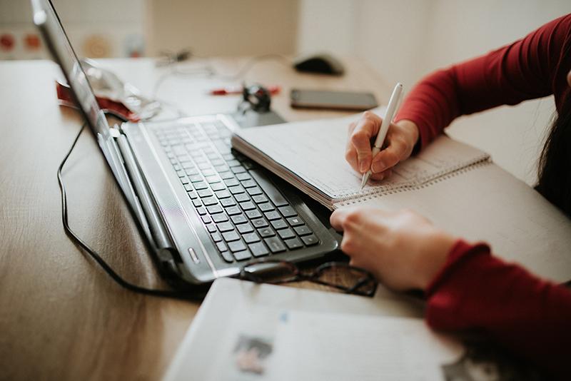 A person sitting at a desk writes in a notebook near a laptop.