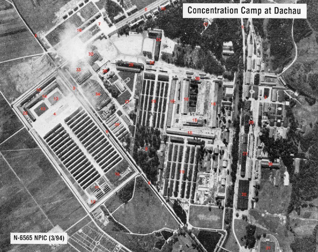 This is a black and white arial photograph titled “Concentration Camp at Dachau.” Another label says, “N-6565 NPIC (3/94)”. In the top middle there is a large group of trees as well as some smaller sections scattered on the right side and in the middle. The left side of the picture shows two organized rows of long, skinny buildings, and an open area. There are roads and sidewalks throughout the picture.