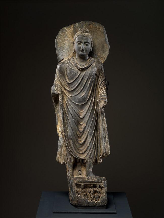 A picture of a golden-black sculpture is shown on a black background. The bare left foot of the man in long flowing robes stands on an ornately carved, square pedestal. His bare right foot is behind him. He is missing his hands and there is a flat circular disc behind his head. He wears a beaded head dressing with a small dome on top of his head, wears earrings, and has a plump, stoic face.