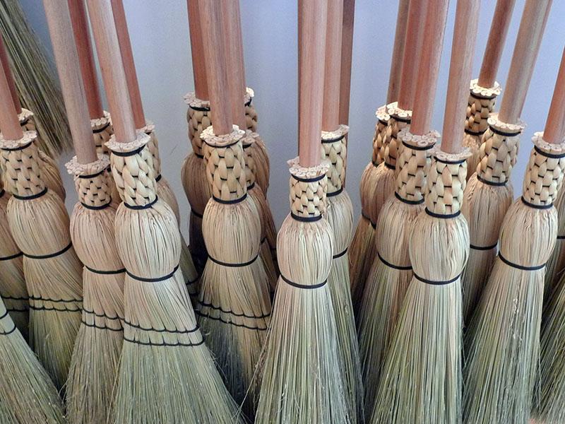 A color photograph of a group of about fifteen straw brooms. The brooms have matching handles of smooth wood. The bottom is constructed of a light-colored plant fiber. The fibers are attached to the brooms using dark twine.