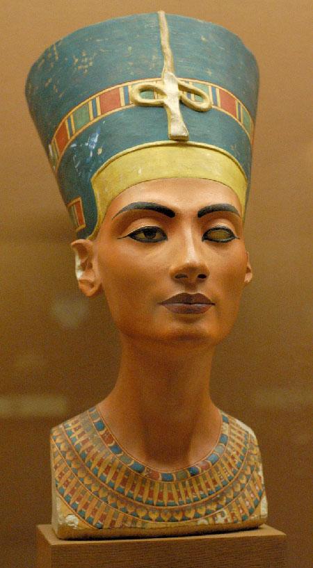 A picture of a colorful bust is shown on a brown background. The bust shows a woman wearing a tall, oval, blue headdress with a gold strip in front and an orange, gold, green and blue decorated stripe running all around the headdress across the middle and behind her right ear. There is a cross-like gold object at the front of the headdress. Her skin is caramel and one eye is black while the other eye is white without a pupil. She has dark eyebrows and maroon full lips. Her right ear is broken off at the top and white shows underneath. She wears a highly colorful necklace in the same colors as her headdress in various shapes and rows. The bottom edges of the bust are chipped and worn.