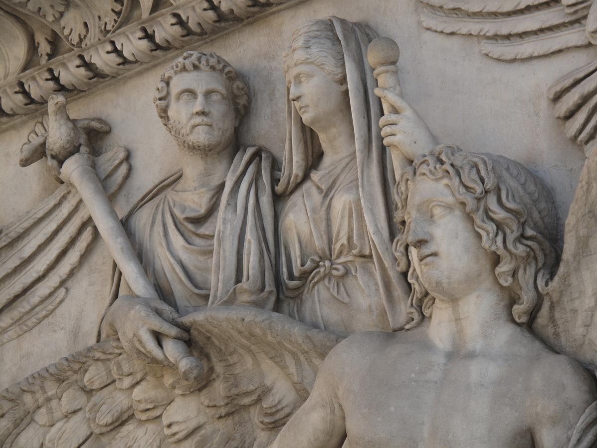 A carving in dirty white stone is shown. The top left corner shows detailed carvings of leaves in the ridges and feathers are carved in the wall on the top right. A man and a woman are carved in the middle. He is shown with short curly hair, a beard and long robes. He holds a long, thick scepter with a winged animal with a curved beak carved at the top. Feathers are carved into the wall behind the scepter. At the right the woman is shown with small eyes, large nose, downturned mouth, wavy hair under a cloth on her head and a dress with a belt tied in the middle. She holds a scepter with a round ball at the top in her left hand. In the bottom right forefront a naked chested figure is shown with shoulder length curly hair and large wings extending out their back. They have almond shaped eyes and a large nose.