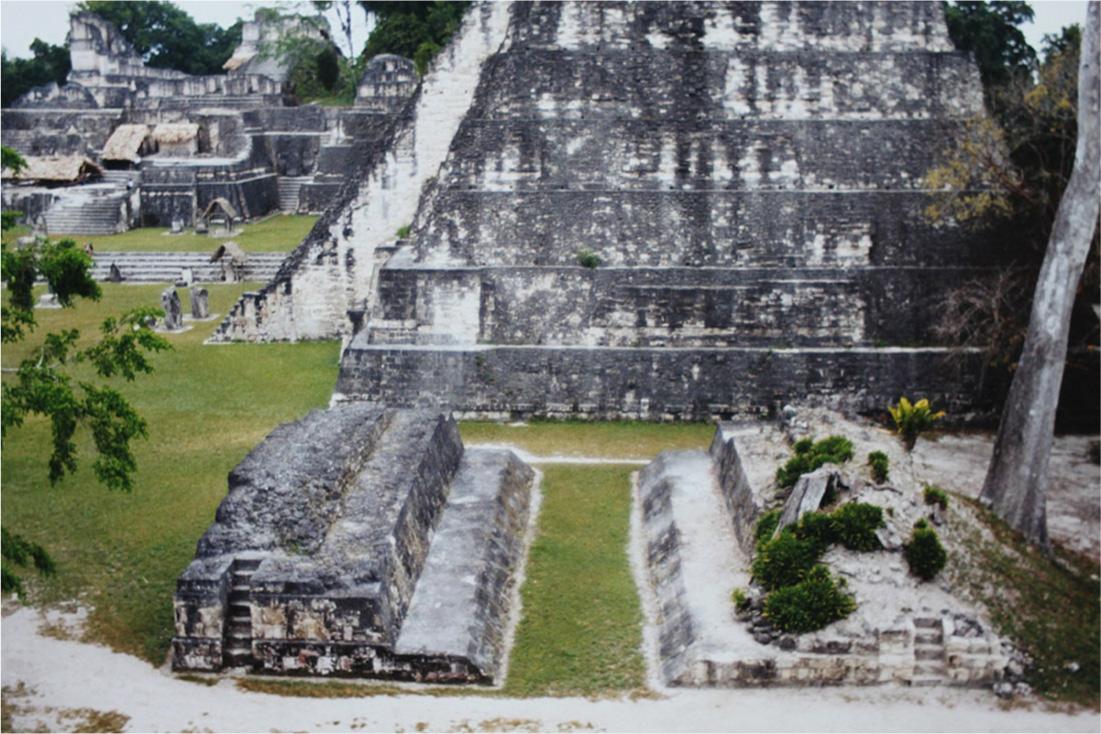 A photo is shown of gray and white stone structures set on green grassy lands with some trees visible at the edges and in the far background. In the top right a large six-tiered pyramid-shaped faded gray and white structure is shown with an angled section running along the left side. In front of the structure two rectangular-shaped gray stone structures lay parallel to each other low to the ground. Stairs can be seen at the front and the top tier is smaller than the bottom tier. A white path is seen running along the front of the photo in front of the two structures. Grasses grow atop the structure on the right as well as a lone tree with no branches. In the top left background small rectangle stones are seen on the ground. Heading into the back left of the photo a wide staircase laid into the ground leads to a flat area with a multi-tiered gray building with some domed and flat structures on the various levels. Many staircases and doorways can be seen. Some brown huts with beige roofs are shown within the structure.