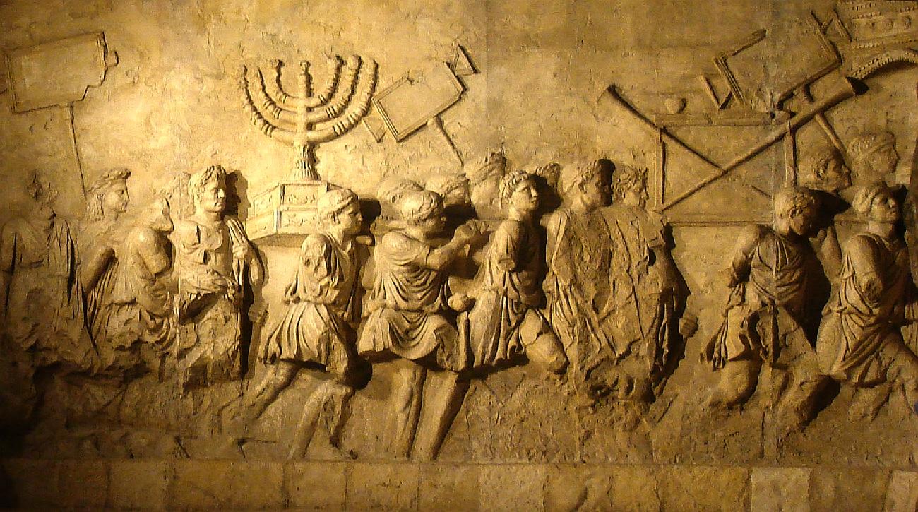 A photograph of an old, faded, and broken in places carving on a wall is shown. Gold colored, the images are carved rising out of the background. The back of the wall is small squares across the bottom and larger squares across the top. In the image, a line of at least seventeen people are walking to the right. All of them wear short robes, belted at the waist, with lariats around their heads. Most have short or shoulder length curly hair and large noses. Many of the legs are missing from the carving as well as some of the figure’s arms and one head. They are seen carrying various items – three rectangular objects on long sticks, a large box with two long sticks in a crisscross pattern across the front, and a large two-tiered and high decorated pedestal with a candle holder with seven arced arms coming out of the middle pole.