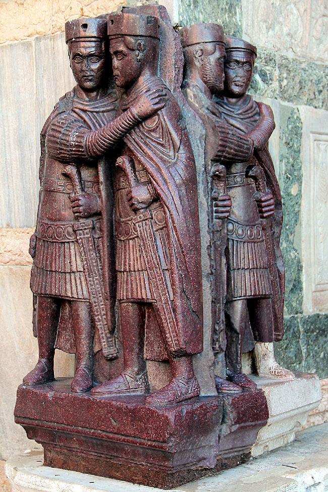 A picture of a stone carving of four men standing together on a short pedestal is shown on a corner where two marble walls meet. The carving is a maroon color and two of the men face left, while two men are behind them facing right. There is a crack running between the two pairs. All four men are shown in dress armor and capes, flat, round hats on their heads, almond shaped eyes, very flat noses, and holding a sword at their sides with their left hands. Both sets of men show the man on the left draping his right arm on the left shoulder of the other man. The left foot of the man furthest in the background as well as his half of the pedestal is all white. The marble wall behind them is various colors of green, yellow, white, and brown.