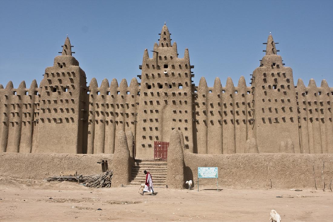 A photograph shows a large, brown structure with many points along the top. Three portions are shown jutting out. Stairs in the front lead to a wooden door. A sign stands at the bottom of the stairs. There is a row of openings at the top of the walls and many projections coming out of the walls in organized rows. A person is walking in front of the stairs on a sandy stretch of land.