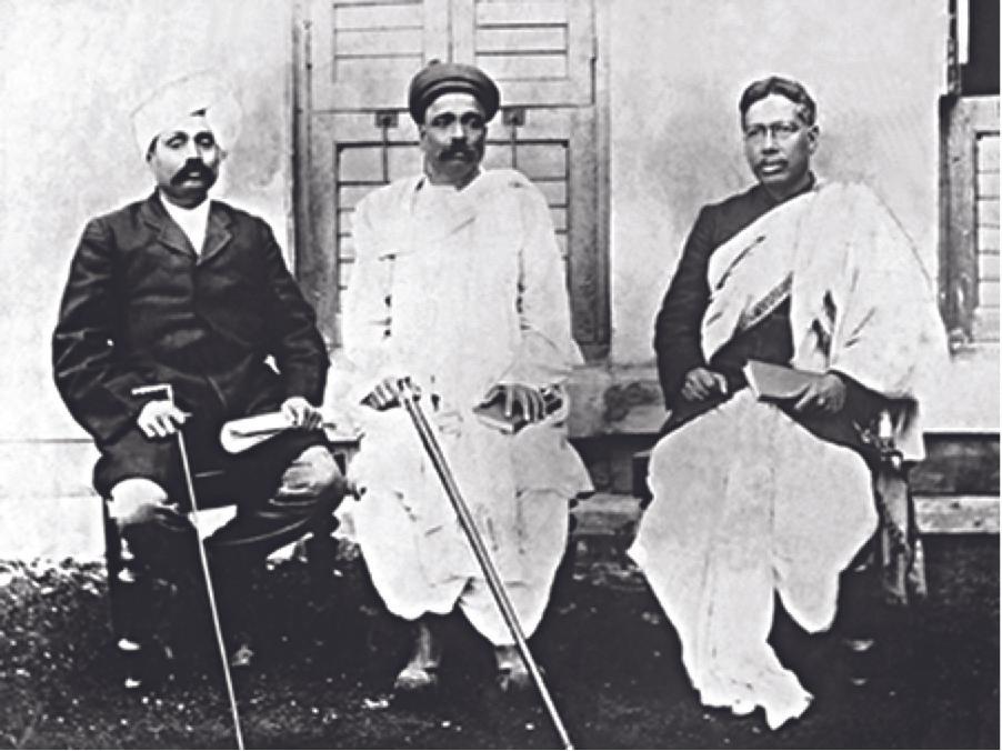 The photograph shows three men seated in chairs in front of a white house with shuttered doors. The man on the left wears a dark, long coat, pants, has a moustache, a white turban on his head and holds a cane in one hand a roll of papers in the other. The man in the middle wears a long white dhoti, has a moustache, a dark small cap, and holds a cane in one hand and a book in the other. The man on the right wears a dark coat and a large white cloth draped over his shoulder and covering his legs. He holds a book in his hand and wears glasses.