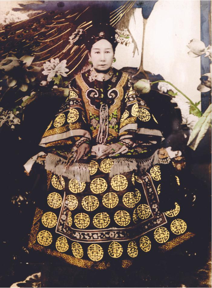 A photo of a woman with a very pale, white face. She wears a long dress decorated with yellow stenciled circles and ornate stitching with very large, flowy sleeves with tassels on the ends. She wears a gold decorated black hat on her head and large earrings. The background shows large flowers.