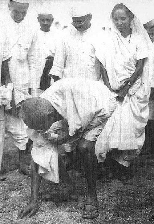 A bald man bends down and picks up some of the earth. Four people stand around him. All are wearing white robes.