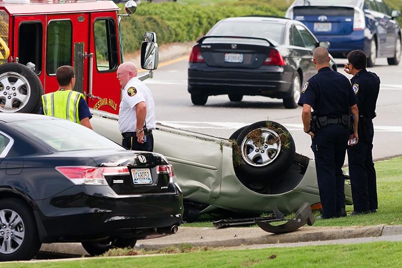 Police officers and first responders stand on a sidewalk next to an overturned car.