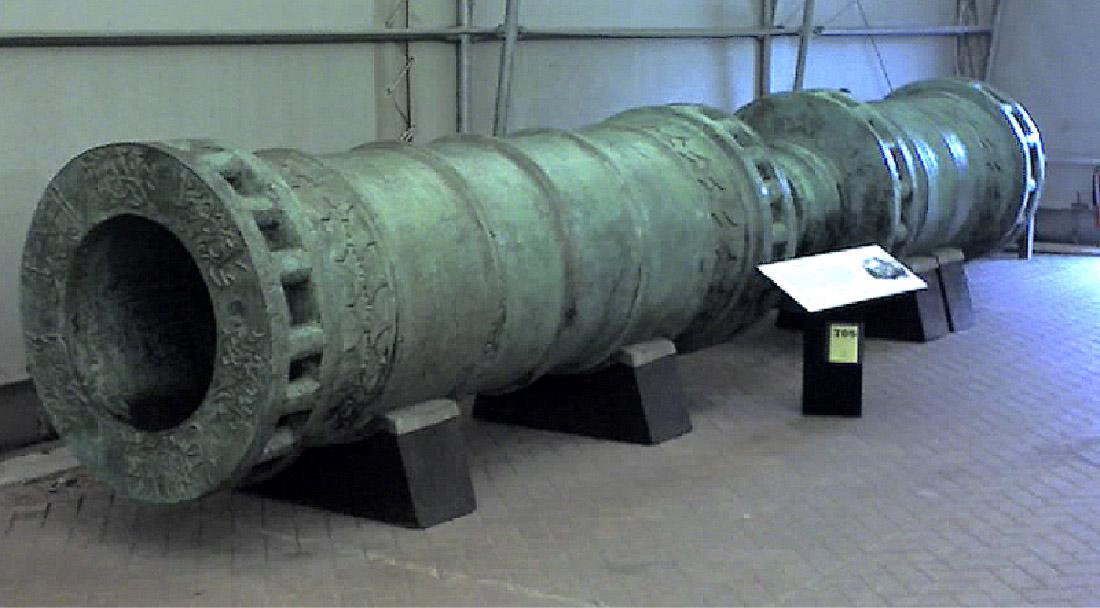 A picture of a long cannon is shown on four black blocks on a brick patterned floor. The wall behind is gray with pipes running up and down and across. The tube is thick and has designs along the bottom opening. Square notches are seen along four segments of the tube. A black podium is seen in front of the tube with a white flat placard at the top.