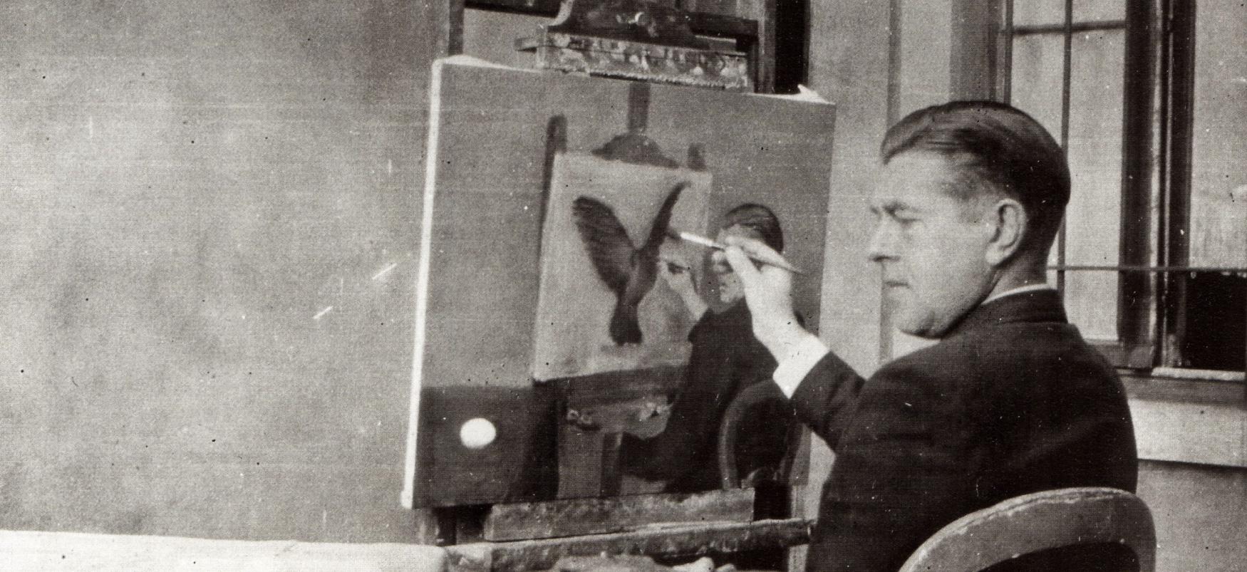 Black and white photograph of a seated man holding a paint brush to a nearly finished canvas resting on an easel.