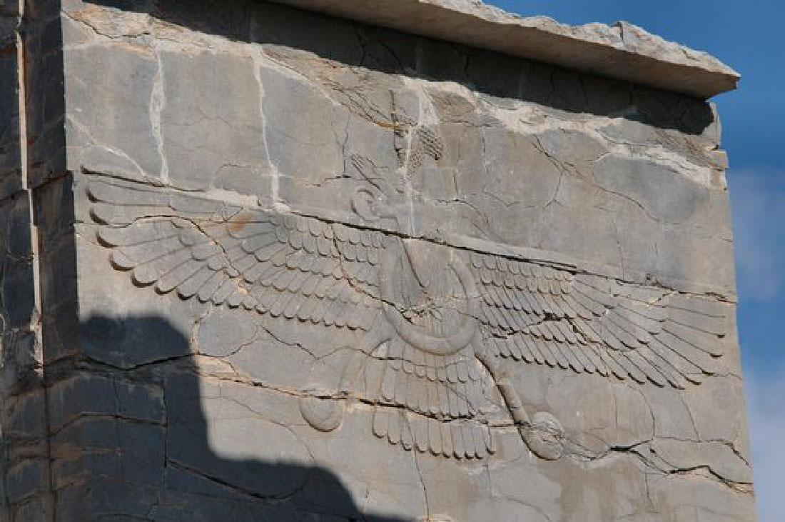 A picture of a square stone carving is shown. At the top is a man’s face facing left with long curly hair and a long curly beard. His right hand is pointed up and his left hand holds a round circular object. He wears long robes that are scalloped below the waist. A large circle is shown below his waist that leads to two large bird legs sticking out at the sides. Large feathered wings spread out from his waist. The stone is cracked and marbled gray and brown. A lip is seen at the top of the stone and a shadow is shown below the stone lip as well as on the left bottom.