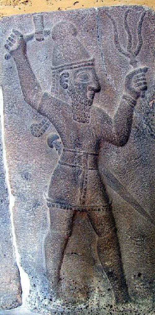 A gray and black stone carving of a figure is shown. A crack runs down one side and the bottom is spotted with black dots. The figure wears a tall hat and has long hair and a long beard. He wears a cloth around his waist with squares along the edges and a sword hangs from his belt. Both of his arms are up in the air and in his right hand he holds an axe. In his left hand he holds a three pronged large item.