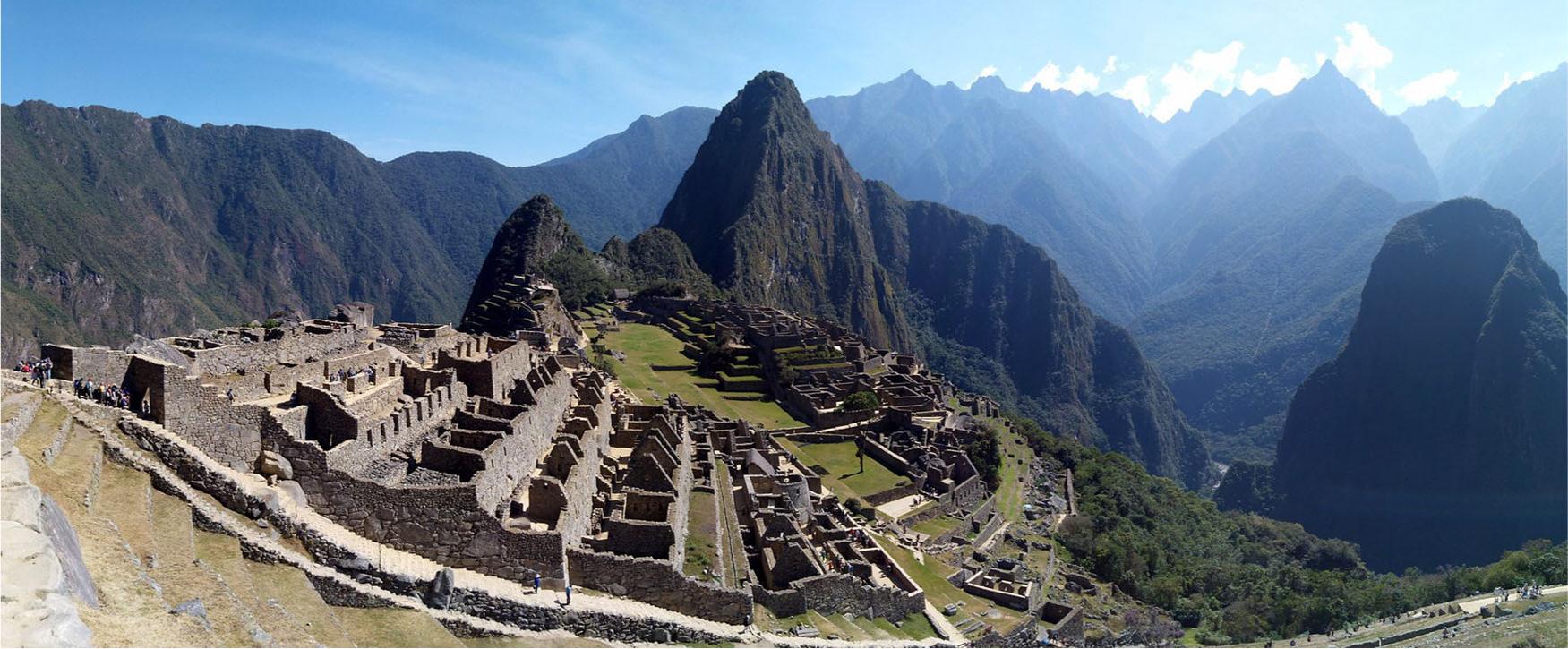 A panoramic photo of ruins of a city on a mountainside with large mountains in the background. In the front left of the image the ruined, dark gray, connected, long stone buildings sit on six steps with walls left depicting rooms and no roofs. Below the last step a flat, green grass filled space shows more stone walls with rooms outlined and roofs missing. Going down the mountain in front of the bottom tier are more ruined stone buildings and a thick forest of green trees. More ruins of maze-like buildings are seen in the background along with a pyramid shaped structure in the back left of the image. The bottom left corner shows beige steps in the mountain with people depicted at the top. In front of those steps are large greenish steps and some rocks. In the bottom right of the image a road can be seen with people standing.